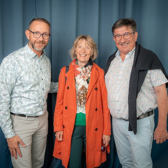 Drei Menschen stehen eng nebeneinander vor einem blauen Vorhang und lächeln. Der Mann links trägt eine Brille und ein gemustertes Hemd. Die Frau in der Mitte trägt einen orangefarbenen Mantel und einen Schal. Der Mann rechts trägt eine schwarze Decke.