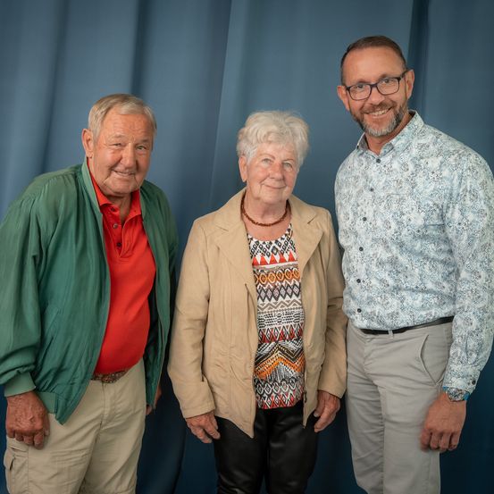Drei Menschen stehen eng beieinander vor einem blauen Vorhang und lächeln für ein Foto. Der Mann links trägt eine grüne Jacke und ein rotes Poloshirt. Die Frau in der Mitte trägt eine beige Jacke und eine Halskette. Der Mann rechts trägt ein gemustertes Hemd und eine Brille.