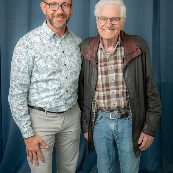 Zwei Männer stehen lächelnd vor einem blauen Vorhang. Der Mann auf der linken Seite trägt ein gemustertes Hemd und graue Hose. Der Mann auf der rechten Seite trägt eine schwarze Lederjacke, ein kariertes Hemd und blaue Jeans.