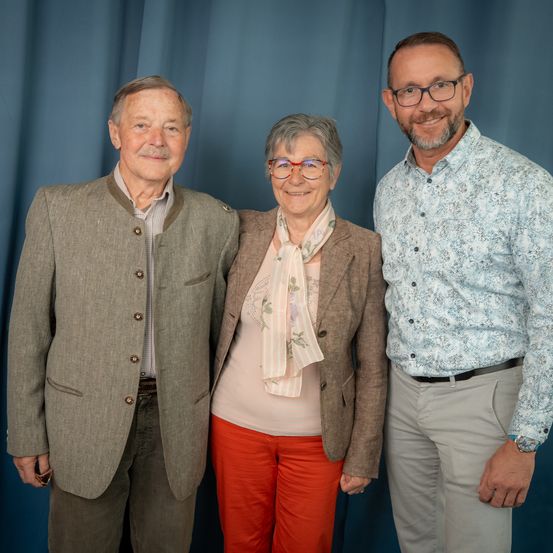 Drei Menschen stehen vor einem blauen Vorhang. Ein Mann links, eine Frau in der Mitte und ein weiterer Mann rechts.