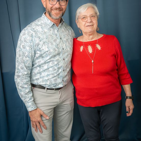 Ein Mann und eine ältere Frau stehen lächelnd eng beieinander vor einem blauen Hintergrund. Der Mann trägt eine Brille, ein gemustertes Hemd und einen Gürtel. Die Frau trägt ein rotes Oberteil, eine Halskette und schwarze Hosen.