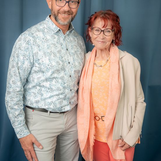 Ein Mann und eine Frau stehen lächelnd nebeneinander vor einem blauen Vorhang. Der Mann trägt ein blau gemustertes Hemd und Khakihose, während die Frau eine beige Jacke, orange Hosen und einen rosa Schal trägt. Beide haben eine Brille auf.