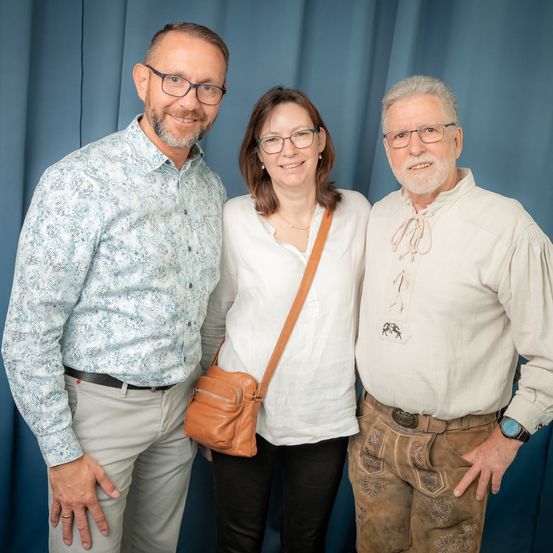 Drei Personen posieren für ein Foto. Ein Mann links trägt ein gemustertes Hemd und eine Brille. Eine Frau in der Mitte trägt eine Halskette und eine Brille. Der Mann rechts trägt ein weißes Hemd und braune Hosen. Sie alle lächeln.