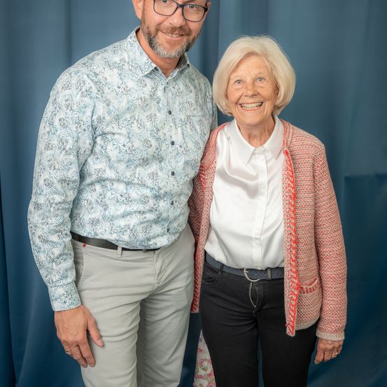 Ein Mann und eine Frau stehen eng nebeneinander und lächeln für ein Foto. Der Mann trägt ein gemustertes Hemd und Khakihosen, während die Frau ein weißes Blusenhemd und eine rosa Strickjacke trägt. Sie stehen vor einem blauen Vorhang.