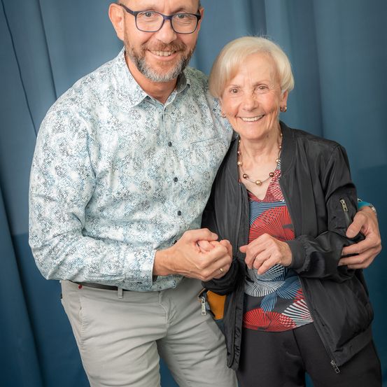 Ein Mann und eine ältere Frau stehen lächelnd nebeneinander und halten sich an den Händen vor einem blauen Vorhang.