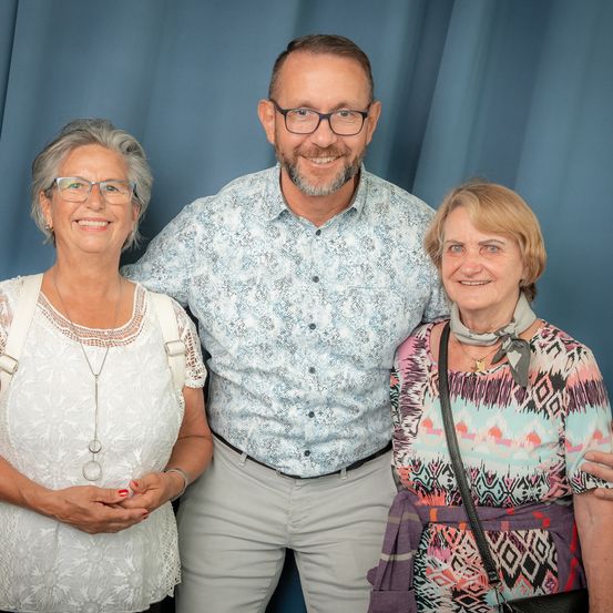 Drei Personen lächeln für ein Foto. Der Mann trägt eine Brille und einen Gürtel, während die Frauen Halsketten, eine Krawatte und eine Tasche tragen. Sie stehen vor einem blauen Vorhang.