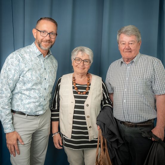 Drei Personen, zwei Männer und eine Frau, stehen eng nebeneinander vor einem blauen Vorhang. Der Mann auf der linken Seite trägt eine Brille und ein Hemd mit Blumenmuster. Die Frau in der Mitte hat eine Halskette und ein gestreiftes Oberteil. Der Mann auf der rechten Seite trägt ein kariertes Hemd und hält eine Jacke.