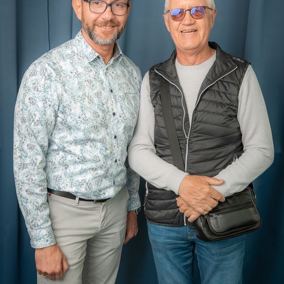 Zwei Männer stehen nebeneinander und lächeln. Der Mann auf der linken Seite trägt ein gemustertes Hemd und einen Gürtel. Der Mann auf der rechten Seite trägt eine Weste und hält eine schwarze Tasche. Beide tragen eine Brille.