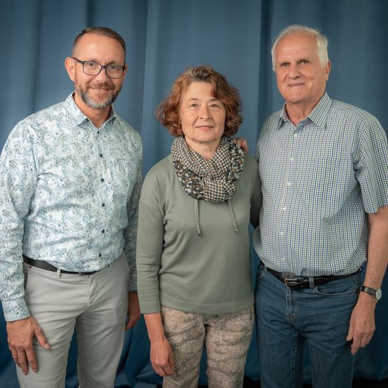 Drei Personen posieren für ein Foto vor einem blauen Vorhang. Der Mann links trägt eine Brille und ein gemustertes Hemd. Die Frau in der Mitte hat eine Schürze und ein grünes Oberteil. Der Mann rechts trägt ein kariertes Hemd und Jeans.