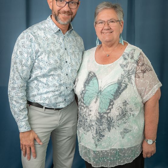 Ein lächelndes Paar steht eng zusammen, mit blauem Hintergrund. Der Mann trägt eine Brille und ein gemustertes Hemd, während die Frau eine Bluse mit Schmetterlingsdruck und Ohrringe trägt.