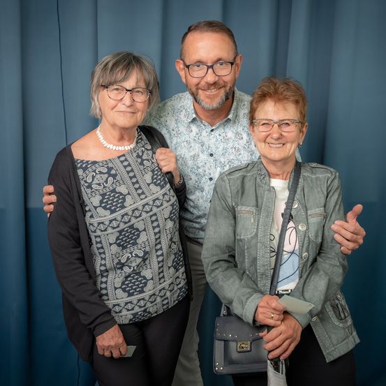 Drei Personen, zwei Frauen und ein Mann, stehen eng beieinander und lächeln für ein Foto vor einem blauen Vorhang.