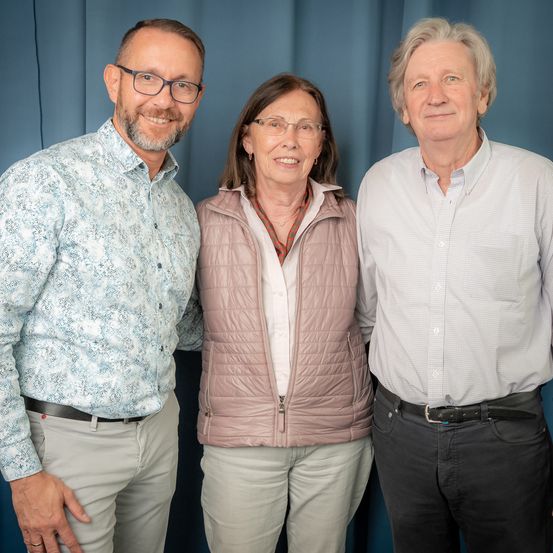 Drei Personen, zwei Männer und eine Frau, stehen eng nebeneinander vor einem blauen Hintergrund und lächeln für die Kamera.