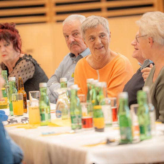 Eine Gruppe älterer Erwachsener sitzt um einen Tisch herum, lächelt und unterhält sich. Der Tisch ist mit verschiedenen Getränken bestückt, darunter Bier und Limonade. Der Raum hat Holzwände und eine Decke.
