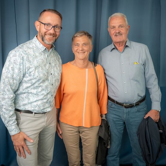 Drei Menschen stehen nebeneinander vor einem blauen Vorhang. Die Frau in der Mitte lächelt.