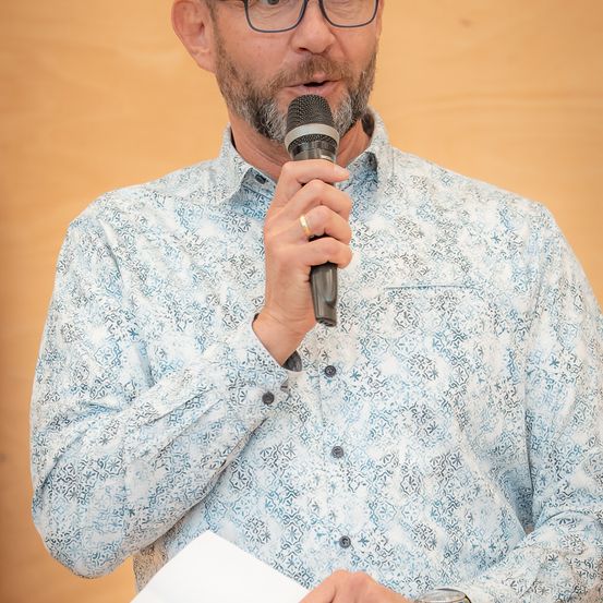 Ein Mann mit Bart hält ein Mikrofon in der rechten Hand und ein Stück Papier in der linken. Er trägt ein gemustertes Hemd und eine Brille.