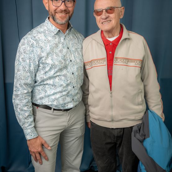 Zwei Männer stehen nebeneinander und lächeln. Der Mann auf der linken Seite trägt ein gemustertes Hemd und graue Hosen. Der Mann auf der rechten Seite trägt eine beige Jacke und hält eine blaue Jacke. Sie stehen vor einem blauen Hintergrund.