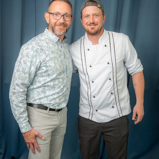 Zwei Männer stehen eng nebeneinander vor einem blauen Vorhang. Einer trägt eine Brille und ein gemustertes Hemd, der andere eine Kochuniform. Beide lächeln.