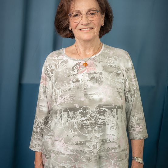 Eine ältere Frau mit kurzen braunen Haaren, die eine Brille und ein graues Blumenhemd mit einer Halskette trägt, lächelt für die Kamera.