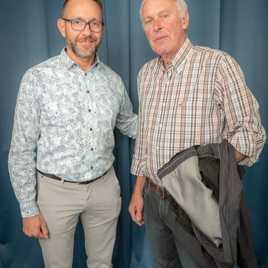 Zwei Männer stehen nebeneinander, einer lächelt und der andere hält eine Jacke.