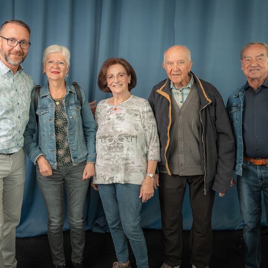 Fünf Personen stehen vor einem blauen Vorhang und scheinen für ein Foto zu posieren.