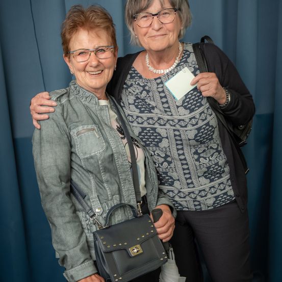 Zwei ältere Frauen stehen eng beieinander und lächeln. Eine trägt eine graue Jacke und eine schwarze Handtasche, die andere ein blau gemustertes Shirt mit Perlenkette. Hinter ihnen ist ein blauer Vorhang.