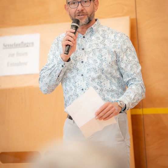 Ein Mann steht mit einem Mikrofon und einem Papier in der Hand und spricht vor einem Publikum. Er trägt ein gemustertes Hemd und eine Hose. Hinter ihm befindet sich eine Holzwand mit einem weißen Schild, auf dem 'Severinlauflagern' und 'nur freund' steht.