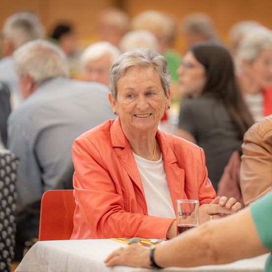 Eine ältere Frau mit grauen Haaren sitzt lächelnd an einem Tisch und trägt eine orangefarbene Jacke und ein weißes Hemd. Ein Glas mit einem Getränk steht vor ihr auf dem Tisch. Hinter ihr sitzen mehrere Personen in einem Raum, wahrscheinlich bei einer gesellschaftlichen Zusammenkunft.