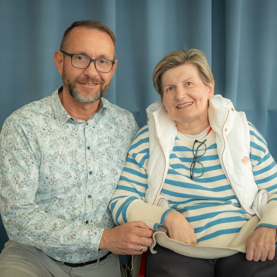 Ein Mann und eine Frau sitzen eng beieinander, beide lächeln, vor einem blauen Hintergrund. Der Mann trägt eine Brille und ein gemustertes Hemd, die Frau eine gestreifte Jacke mit einer Brille daran.