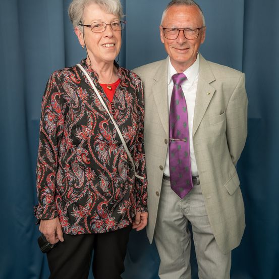 Ein älteres Paar steht nebeneinander mit Lächeln im Gesicht. Die Frau trägt eine Brille, eine Paisley-Bluse und schwarze Hose. Der Mann ist in einem Anzug und einer lila Krawatte gekleidet. Sie stehen vor einem blauen Vorhang.