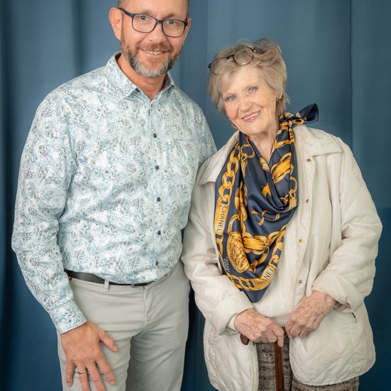 Ein Mann und eine Frau stehen nebeneinander. Der Mann trägt eine Brille und ein gemustertes Hemd. Die Frau hat eine Schürze um den Hals und hält einen Stock. Sie lächeln beide und scheinen für ein Foto zu posieren.