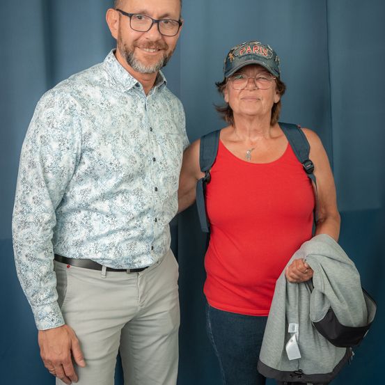 Ein Mann und eine Frau stehen nebeneinander, beide tragen eine Brille. Der Mann ist in einem gemusterten Hemd und Hose gekleidet. Die Frau trägt ein rotes Tanktop, Jeans und einen Paris-Hut. Sie hält eine graue Jacke in der Hand.