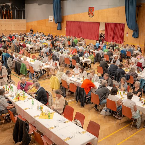 Eine große Gruppe von Menschen sitzt an langen Tischen in einem Saal, wahrscheinlich bei einer Gemeinschaftsveranstaltung. Rote Vorhänge und blaue Draperien sind im Hintergrund.