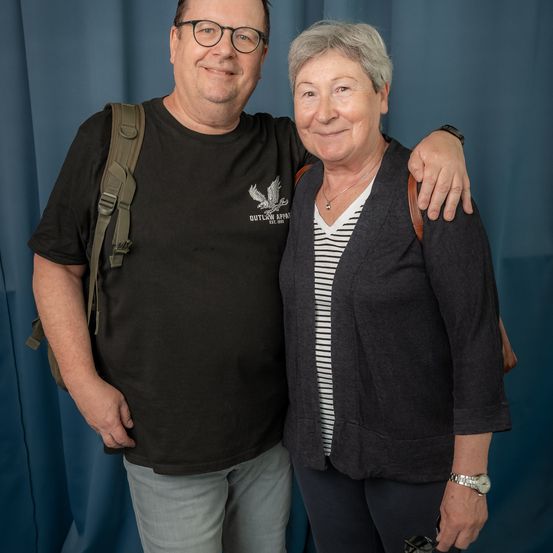 Ein Mann mit Brille und Rucksack posiert mit einer lächelnden Frau, beide stehen vor einem blauen Vorhang.