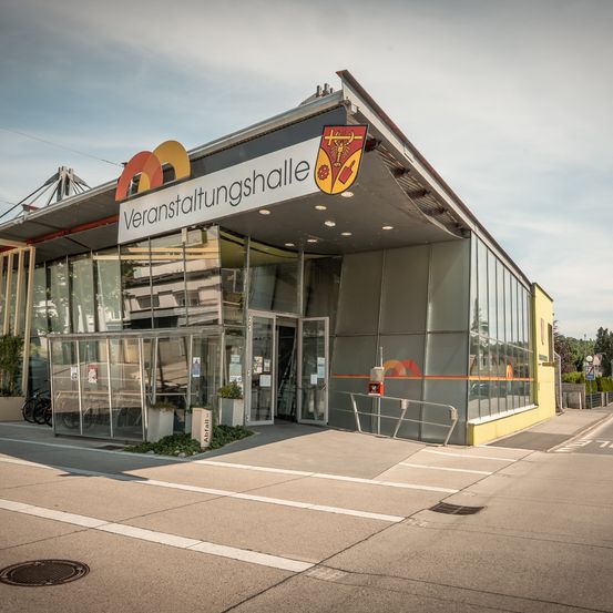 Ein leeres Gebäude außen mit Glaswänden und einem Schild mit der Aufschrift 'Veranstaltungshalle' ist zu sehen. Das Gebäude hat ein modernes Design mit einem Glastüreingang und vor dem Gebäude geparkten Fahrrädern.