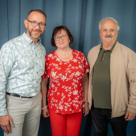 Drei Menschen stehen zusammen vor einem blauen Vorhang. Der Mann links trägt eine Brille und ein Blumenhemd. Die Frau in der Mitte trägt ein rotes Blumenhemd und Hose. Der Mann rechts trägt ein grünes Hemd und eine beige Jacke.