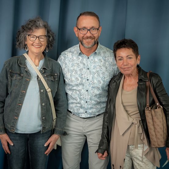 Drei Menschen stehen eng beieinander vor einem blauen Vorhang und lächeln für ein Foto. Der Mann trägt eine Brille und ein Blumenhemd, die beiden Frauen haben kurze Haare und tragen Jeans- und Lederjacken.