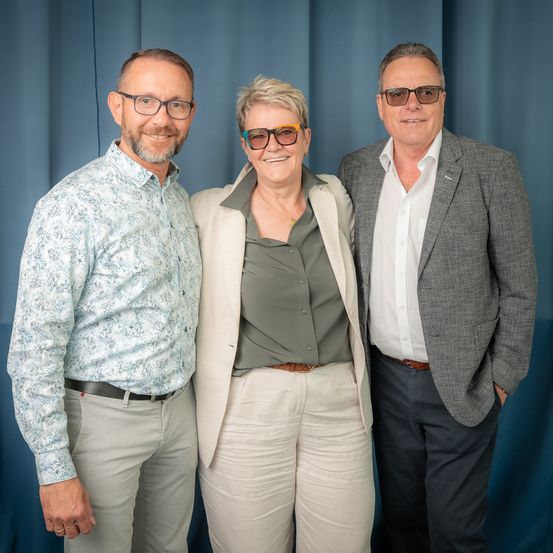 Drei Personen stehen eng beieinander und lächeln für ein Foto. Ein Mann auf der linken Seite trägt eine Brille und ein gemustertes Hemd. Die Frau in der Mitte trägt eine Brille und eine Halskette. Der Mann auf der rechten Seite trägt einen Blazer und ein weißes Hemd.