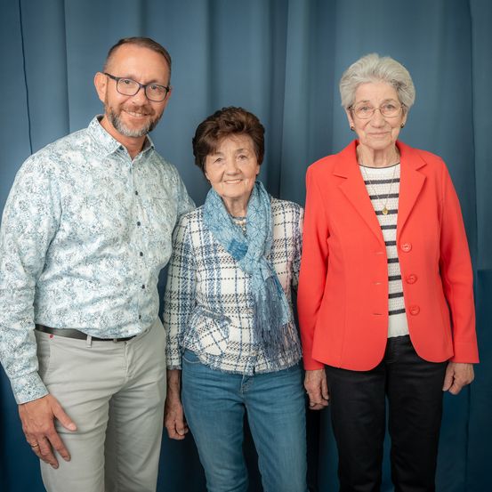 Drei Menschen stehen eng nebeneinander vor einem blauen Vorhang. Ein Mann trägt eine Brille und ein Hemd mit Blumenmuster und lächelt. Daneben steht eine Frau in einer karierten Bluse und einem blauen Schal. Rechts davon eine ältere Frau in einem roten Blazer und einer Brille, die lächelt.