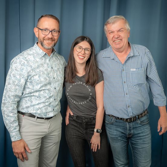 Drei Personen posieren für ein Foto. Der Mann links trägt ein gemustertes Hemd, die Frau in der Mitte ein graues T-Shirt mit Tom Taylor Denim und der Mann rechts ein blau kariertes Hemd. Sie alle lächeln.