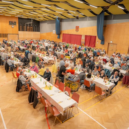 Eine große Gruppe von Menschen sitzt an Tischen in einer Halle, einige mit Getränken. Der Raum ist gut beleuchtet, mit Holzböden und blauen Vorhängen. Die Wände sind in einer warmen Farbe gestrichen.