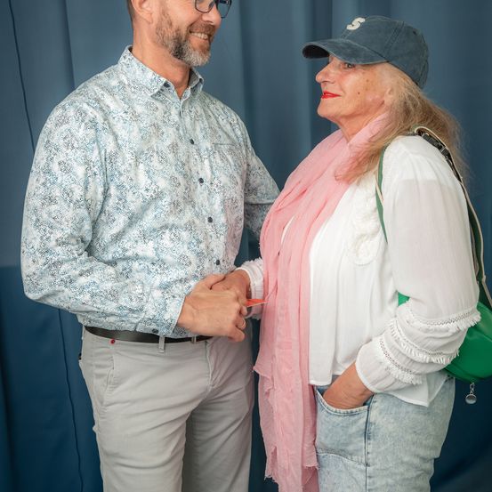 Ein Mann und eine Frau stehen lächelnd nebeneinander. Der Mann trägt eine Brille und ein gemustertes Hemd, während die Frau eine Kappe und einen rosa Schal trägt. Sie halten sich an den Händen, der Mann in formeller Kleidung und die Frau mit einer grünen Tasche.