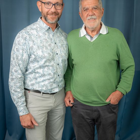 Zwei Männer stehen eng beieinander und lächeln für ein Foto. Der Mann auf der linken Seite trägt ein gemustertes Hemd und graue Hose. Der Mann auf der rechten Seite trägt einen grünen Pullover und dunkle Hose.
