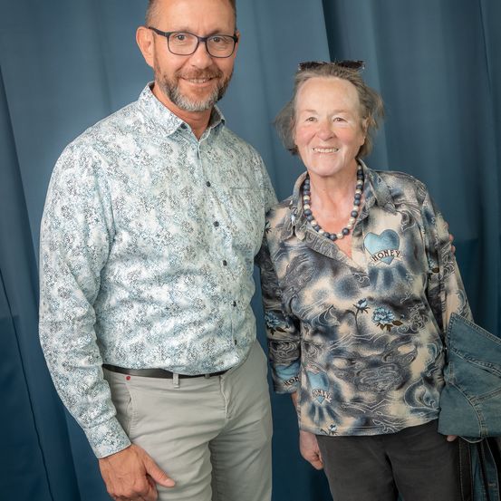 Ein Mann und eine Frau stehen nebeneinander und lächeln vor einem blauen Vorhang. Der Mann trägt ein gemustertes Hemd und eine Brille, die Frau ein Bluse mit Blumenmuster und einem Herz sowie eine Perlenkette.