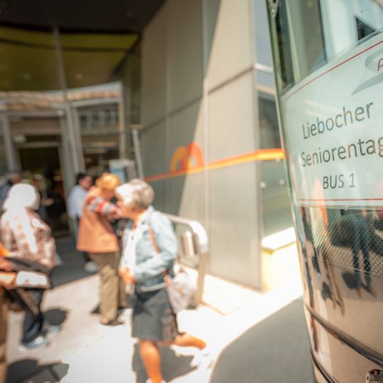 Ein Bus mit der Aufschrift 'Liebocher Seniorentag BUS 1' steht geparkt, während Menschen auf dem Bürgersteig gehen. Einige Menschen sind verschwommen.