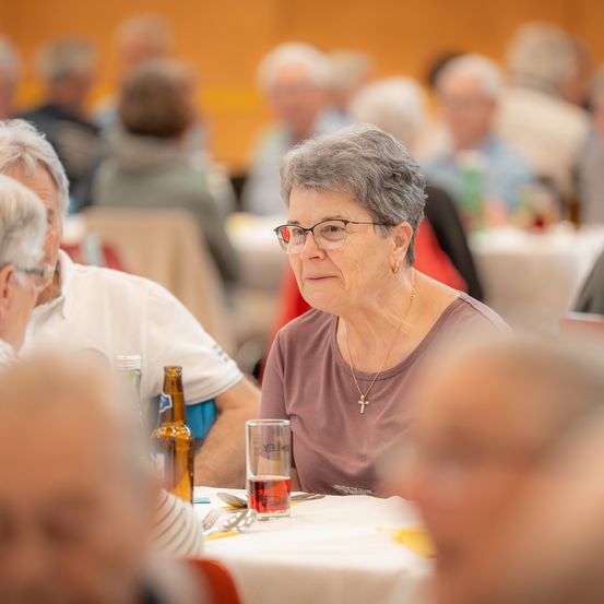 Eine ältere Frau mit Brille und einer Halskette sitzt an einem Tisch mit einem Glas roter Flüssigkeit. In der Nähe sitzt ein Mann neben ihr. Sie befinden sich in einem verschwommenen Hintergrund von essenden Menschen.