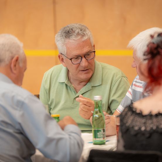 Eine Gruppe älterer Menschen sitzt um einen Tisch. Ein Mann redet, während die anderen zuhören. Flaschen und Gläser stehen auf dem Tisch.