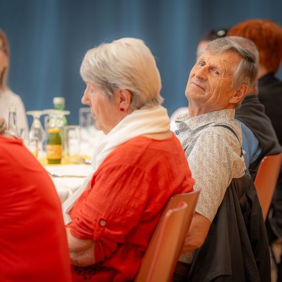 Zwei ältere Personen sitzen in Stühlen, möglicherweise in einem Restaurant. Die Frau trägt ein rotes Oberteil und einen weißen Schal. Der Mann trägt ein Blumenhemd und lächelt.