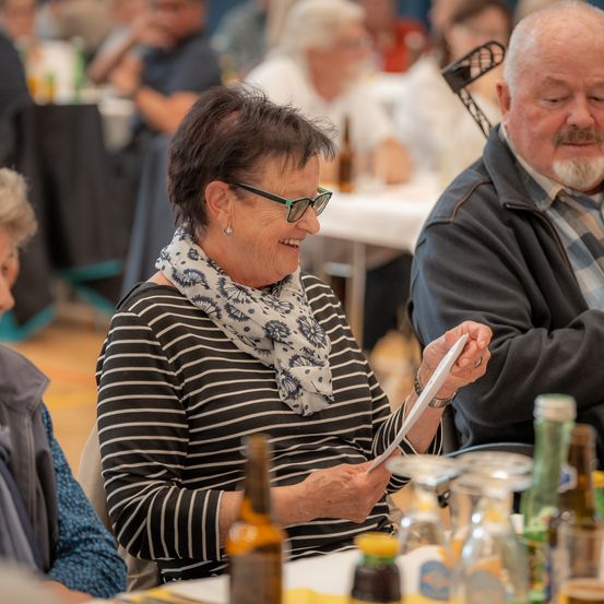 Eine ältere Frau in einem gestreiften Shirt lacht, während sie ein Stück Papier in der Hand hält. Neben ihr lächelt ein älterer Mann. Sie sind umgeben von Flaschen, Gläsern und einem Tisch. Im Hintergrund sitzen andere Menschen an Tischen.