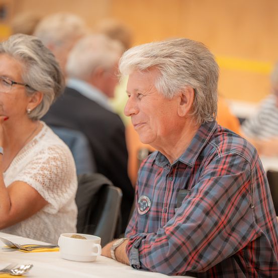 Ein älterer Mann und eine ältere Frau sitzen an einem Tisch. Der Mann trägt ein kariertes Hemd und hat die Augen geschlossen. Die Frau trägt eine Brille und hat die Hand am Kinn. Hinter ihnen sitzen andere an Tischen.