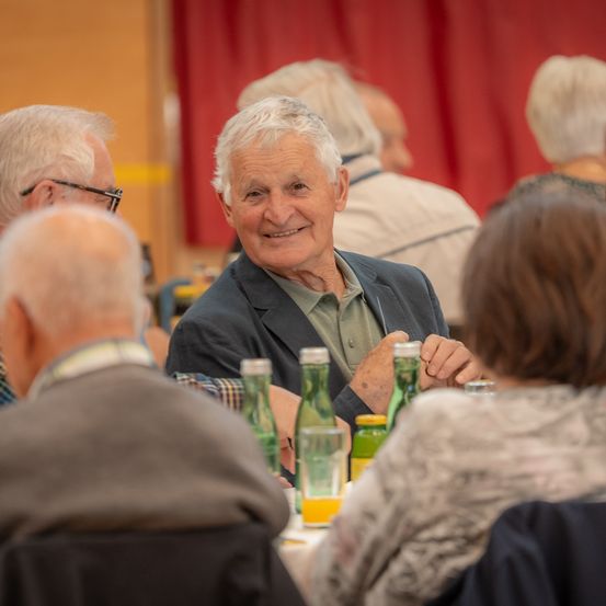 Ein älterer Mann sitzt lächelnd am Tisch, umgeben von anderen älteren Personen, wahrscheinlich bei einer Zusammenkunft oder Versammlung. Flaschen und Gläser stehen auf dem Tisch.